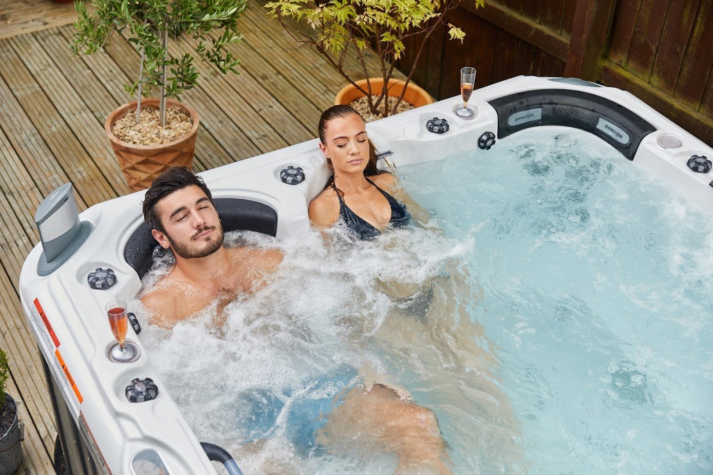 Two people relaxing in a hot tub on a wooden deck with plants around.