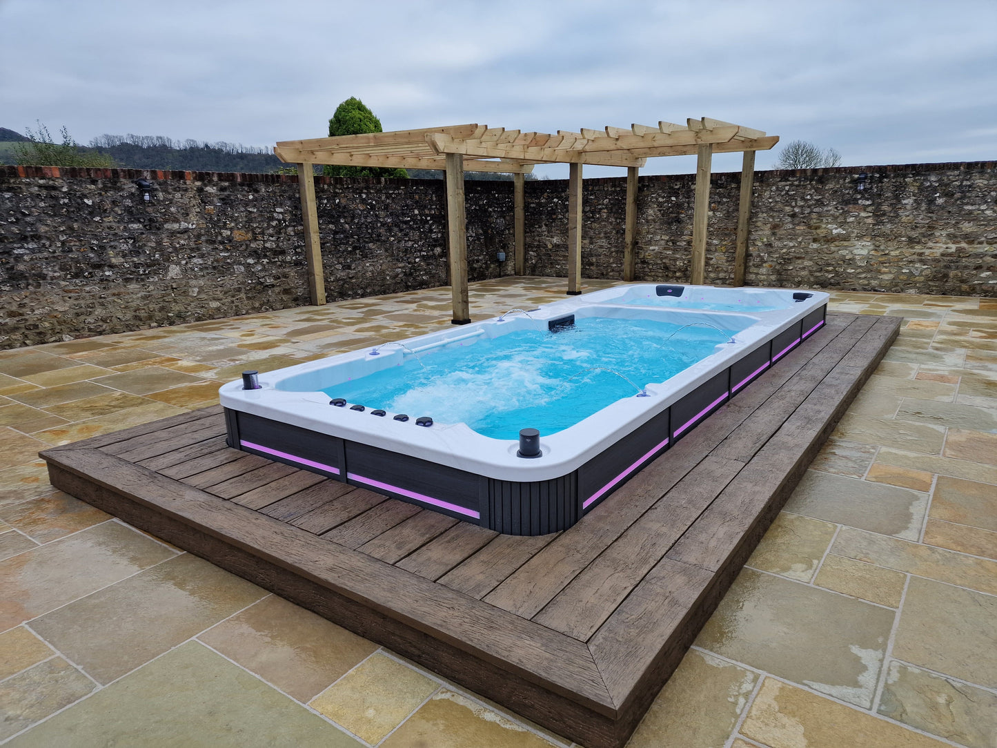 Inground dual temperature swimspa on a wooden deck with a stone wall and pergola in the background